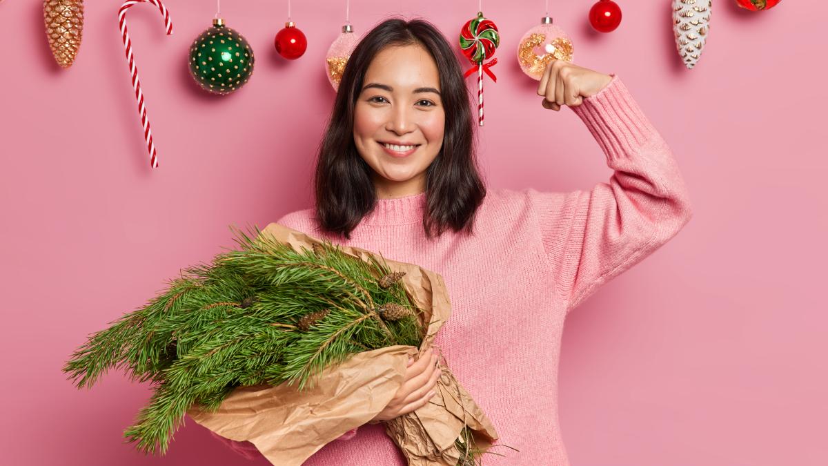 happy-mixed-race-woman-holds-bouquet-fir-tree-branches-raises-arm-shows-how-strong-she-is-dressed-sweater-poses