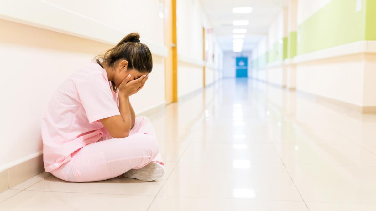 stressed-young-nurse-covering-face-while-sitting-floor-corridor-hospital