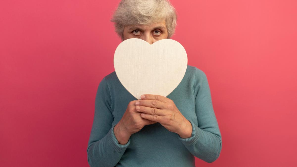 old-woman-wearing-blue-turtleneck-sweater-holding-heart-shape-looking-front-from-it-isolated-pink-wall